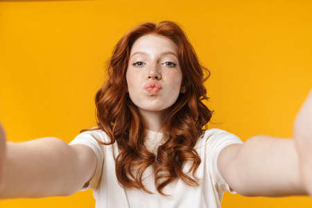 Image of redhead cheerful girl making kiss lips and taking selfie photo isolated over yellow backgroundの写真素材