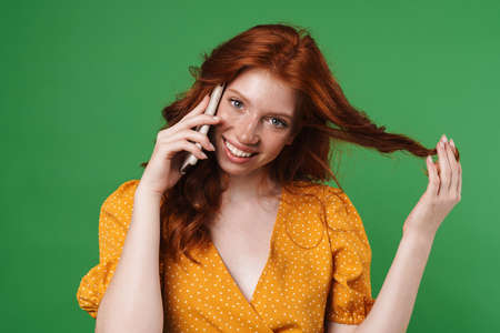 Image of ginger happy girl smiling and talking on mobile phone isolated over green backgroundの写真素材