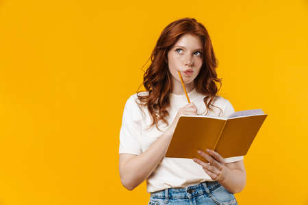 Image of ginger serious girl thinking and holding exercise book isolated over yellow background