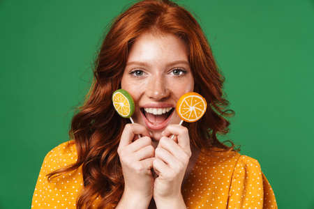 Image of redhead funny girl smiling and holding lollipops isolated over green backgroundの写真素材