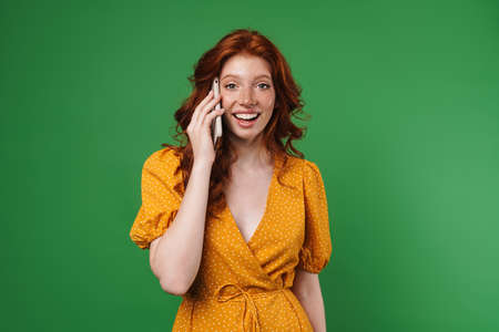 Image of ginger happy girl smiling and talking on mobile phone isolated over green backgroundの写真素材