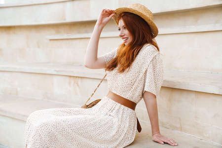 Image of young ginger girl in straw hat smiling while sitting on stairs at summer day