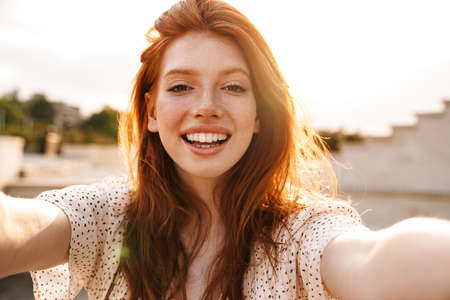 Image of cheerful ginger girl taking selfie and smiling at camera at city street