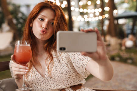 Image of pleased ginger girl taking selfie on mobile phone and drinking cocktail while sitting in...