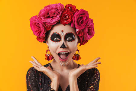 Delighted girl in halloween makeup and flower wreath posing at camera isolated over yellow wall