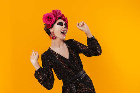 Joyful girl in halloween makeup and flower wreath laughing on camera isolated over yellow wall