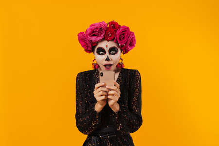 Shocked girl in halloween makeup and flower wreath using mobile phone isolated over yellow wallの写真素材
