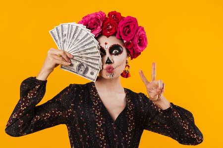 Amusing girl in halloween makeup gesturing peace sign while holding dollars isolated over yellow wallの写真素材