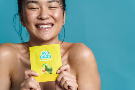 Beauty portrait of a smiling topless young asian woman posing while standing isolated over blue background, showing avocado moisturizerの写真素材
