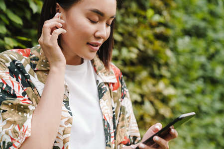 Attractive young asian woman listening to music with wireless earphones while standing near a green bush, holding mobile phoneの写真素材
