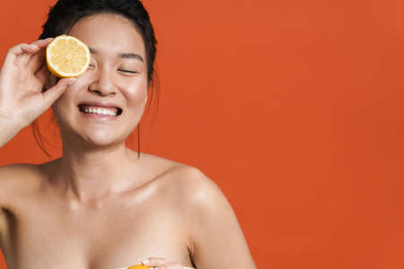 Image of shirtless asian girl laughing while posing with lemon isolated over orange backgroundの写真素材