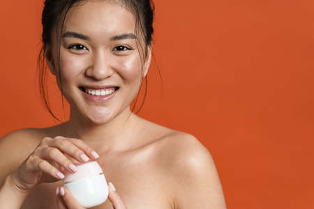Image of smiling shirtless asian girl posing with facial cream isolated over orange backgroundの写真素材