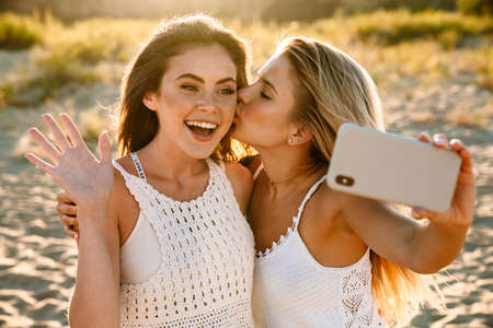Two young caucasian happy women smiling and taking selfie photo at cellphone while walking outdoorsの写真素材