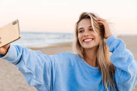 Young caucasian blonde woman smiling and taking selfie photo on cellphone outdoorsの写真素材