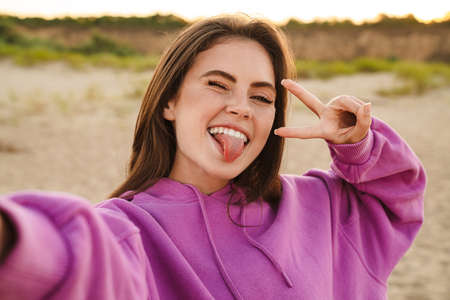 Young caucasian beautiful woman gesturing peace sign and taking selfie photo outdoors