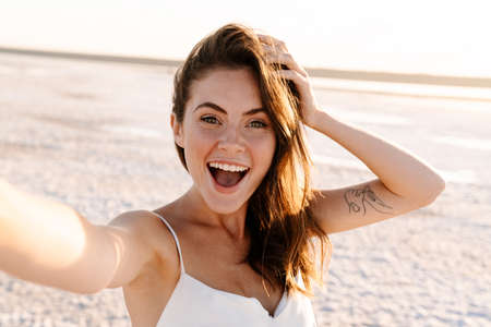 Picture of a cute surprised excited young pretty girl walking outdoors at the beach and taking a selfie by cameraの写真素材