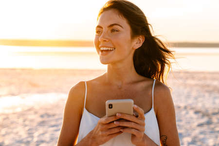 Image of a smiling young pretty girl walking outdoors at the beach and using mobile phoneの写真素材