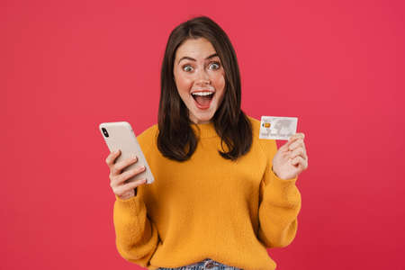 Image of delighted caucasian girl posing with cellphone and credit card isolated over pink...