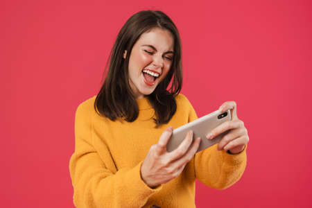Image of excited brunette girl playing video game on cellphone isolated over pink background