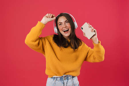 Happy young beautiful woman isolated over pink wall background, listening to music with headphones, using mobile phoneの写真素材