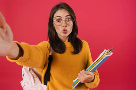Image of surprised student girl with exercise books taking selfie photo isolated over pink backgroundの写真素材