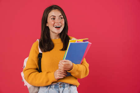Cheerful young girl student wearing backpack, carrying textbooks standing isolated over pink backgroundの写真素材