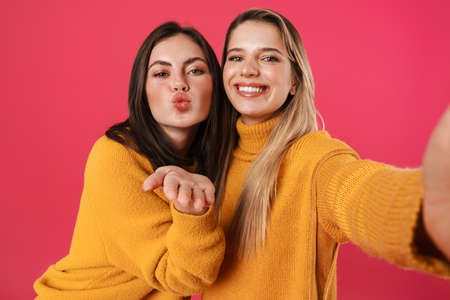 Image of happy beautiful women blowing air kiss while taking selfie isolated over pink background