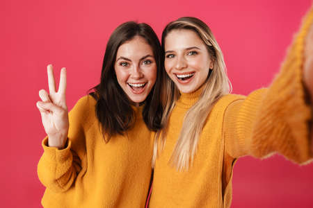 Image of smiling beautiful women showing peace sign while taking selfie isolated over pink backgroundの写真素材