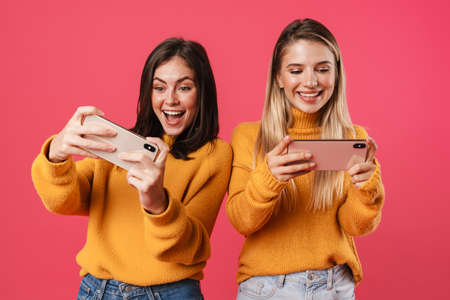 Image of delighted nice women playing video game on smartphones isolated over pink backgroundの写真素材