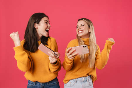 Two happy pretty girls celebrating success while using mobile phones isolated over pink backgroundの写真素材