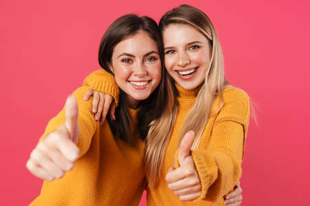 Portrait of two happy young female friends standing over pink background, showing thumbs upの写真素材