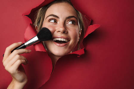 Caucasian young beautiful brunette woman applying makeup with brush through red background isolated closeupの写真素材
