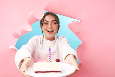 Caucasian young attractive woman smiling and holding birthday cake through pink background isolatedの写真素材