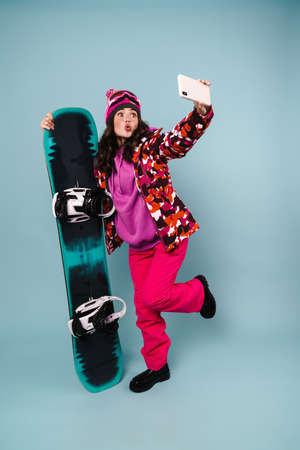 Amusing sportswoman taking selfie on cellphone while posing with snowboard isolated over blue wallの写真素材