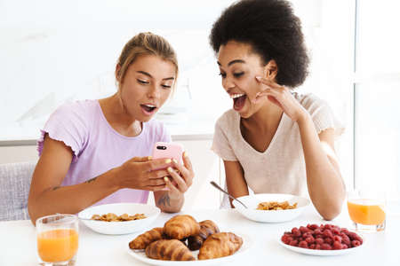 Nice surprised multicultural girls using cellphone while having breakfast together at home kitchenの写真素材