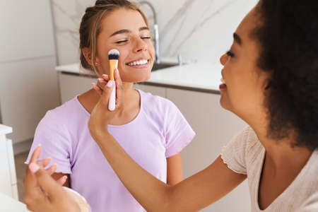 Beautiful joyful multicultural girls smiling while doing makeup together at home kitchen