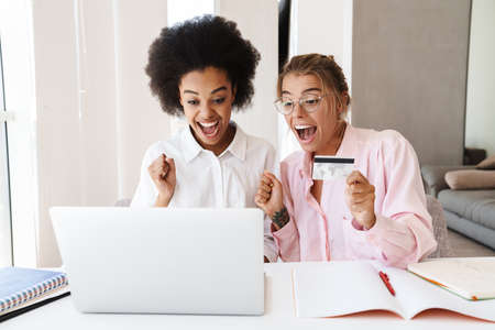 Excited multicultural students girls making winner gesture while using credit card and laptop together at homeの写真素材