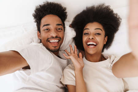 Beautiful happy young african couple relaxing in bed, taking a selfieの写真素材