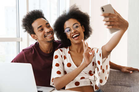 Portrait of a beautiful happy couple sitting at table with laptop computer on a kitchen at home and taking a selfieの写真素材