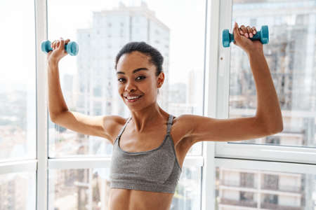 Photo of a young happy cheerful sports african woman making exercises with dumbbells indoors