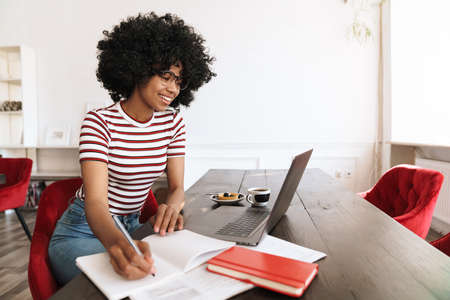 Smiling african american student girl doing homework with laptop while sitting in cafe