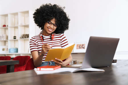 Smiling african american student girl doing homework with laptop while sitting at table indoors