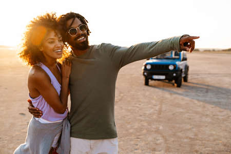 Image of happy african american couple pointing finger and walking while travelling with car on...