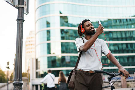 Cheerful african american guy in headphones using cellphone while standing with bicycle at city streetの写真素材