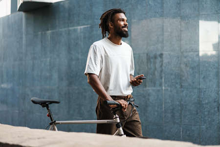 Smiling african american guy using cellphone while walking with bicycle at city streetの写真素材
