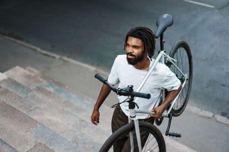 Pleased african american guy smiling while walking bicycle on his shoulder at city streetの写真素材