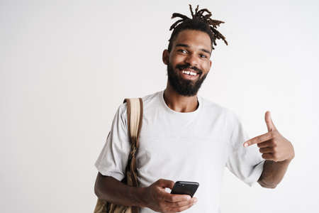 Photo of joyful african american guy holding and pointing finger at cellphone isolated over white backgroundの写真素材