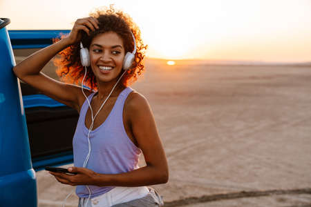 Image of happy african american woman using headphones and cellphone while travelling with car on...