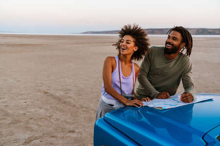 Image of joyful african american couple examining map while travelling with car on desert