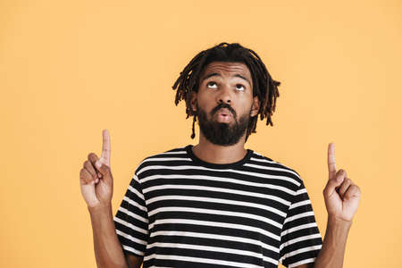 Photo of confused african american guy pointing fingers upward isolated over yellow background